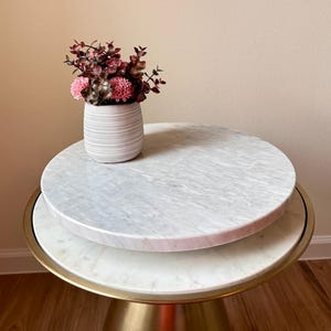 May include: A decorative side table with a white marble top and a gold base. A white, ribbed vase holds pink and burgundy flowers. The table has a round shape and is suitable for a living room or bedroom.