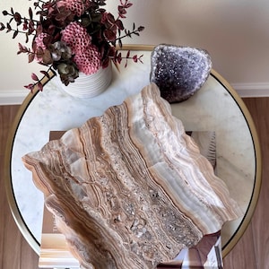 May include: A square tray made of banded brown and white onyx stone with a wavy, scalloped edge. The tray features natural horizontal striations in shades of tan, cream, and dark brown, showcasing the unique mineral layers of the polished stone.