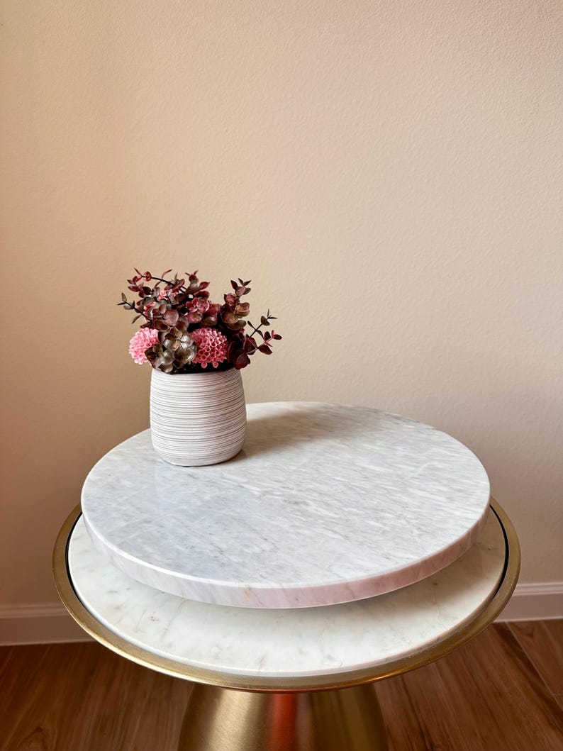 May include: A decorative side table with a white marble top and a gold base. A white, ribbed vase holds pink and burgundy flowers. The table has a round shape and is suitable for a living room or bedroom.
