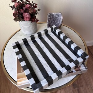 May include: A square decorative tray featuring a bold, alternating black and white striped pattern. The tray is constructed with a smooth, polished finish and has raised edges that continue the striped design around the entire perimeter.