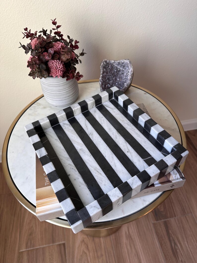 May include: A square decorative tray featuring a bold, alternating black and white striped pattern. The tray is constructed with a smooth, polished finish and has raised edges that continue the striped design around the entire perimeter.