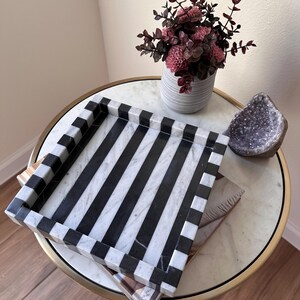 May include: A square tray featuring a bold black and white striped pattern. The tray has raised edges with a matching alternating black and white block design. The surface of the tray is smooth and flat, showcasing a high-contrast geometric aesthetic.