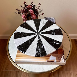 May include: A round, black and white marble decorative piece with a radial design sits atop a book titled "The Soft Minimalist Home" on a gold-rimmed table. A vase of pink and burgundy flowers and a crystal are in the background.