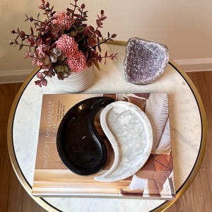 Puede incluir: Una composición decorativa sobre una mesa redonda. El arreglo incluye una bandeja en forma de yin yang en blanco y negro, una geoda y un jarrón con flores rosas. La mesa tiene una tapa de mármol y un borde dorado.