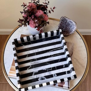 Op de afbeelding: Een vierkant decoratief dienblad met een zwart-wit streepjespatroon. De lade is gemaakt van marmer en staat op een tafel met marmeren blad. Een vaas met roze bloemen en een paarse geode staan ook op de tafel.