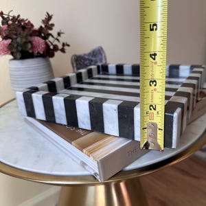 May include: A rectangular tray featuring a bold black and white striped pattern made of stone. A yellow Stanley measuring tape is positioned vertically against the side of the tray, indicating a height of approximately 2.5 inches for the rim.