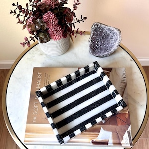 May include: A decorative tray with black and white stripes sits on a table with a marble top. A book titled "The Soft Minimalist" is beneath the tray. A small white vase with pink flowers and a geode are also on the table.