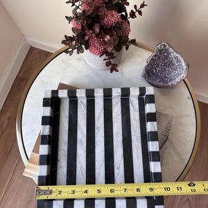 May include: A rectangular tray featuring alternating black and white marble stripes. A yellow Stanley measuring tape is placed across the front edge, indicating a width of approximately 12 inches. The tray has raised edges and a smooth, polished finish.