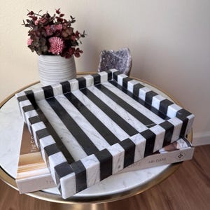 May include: A square decorative tray featuring a bold, alternating black and white striped pattern. The tray is constructed with a smooth, polished finish and has raised edges, showcasing a high-contrast geometric design throughout the entire surface.