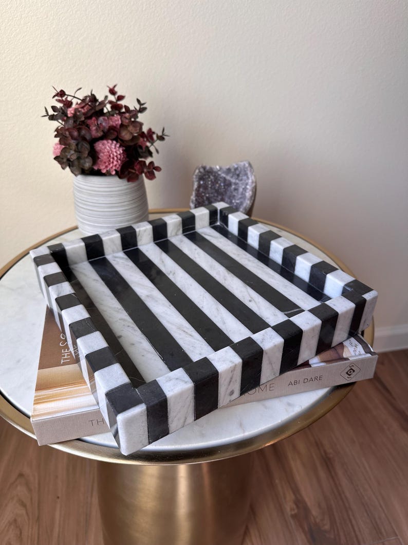 May include: A square decorative tray featuring a bold, alternating black and white striped pattern. The tray is constructed with a smooth, polished finish and has raised edges, showcasing a high-contrast geometric design throughout the entire surface.