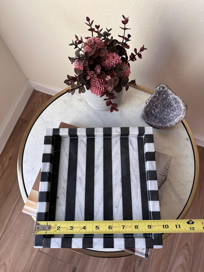May include: A rectangular marble tray featuring a bold black and white vertical striped pattern. A yellow Stanley measuring tape is positioned across the front edge, indicating a width of approximately 11.5 inches for the tray's interior base.