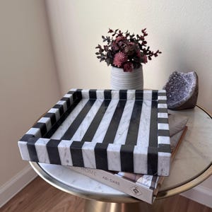 May include: A square tray featuring a bold black and white striped pattern. The tray is constructed with thick, raised edges and a flat base, showcasing a smooth, polished stone-like texture with alternating dark and light vertical stripes.
