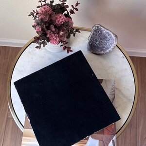 May include: A square black velvet display board resting on a book, accompanied by a white ribbed vase containing pink flowers with dark foliage and a jagged purple amethyst crystal geode, all arranged on a round white marble table with a gold rim.
