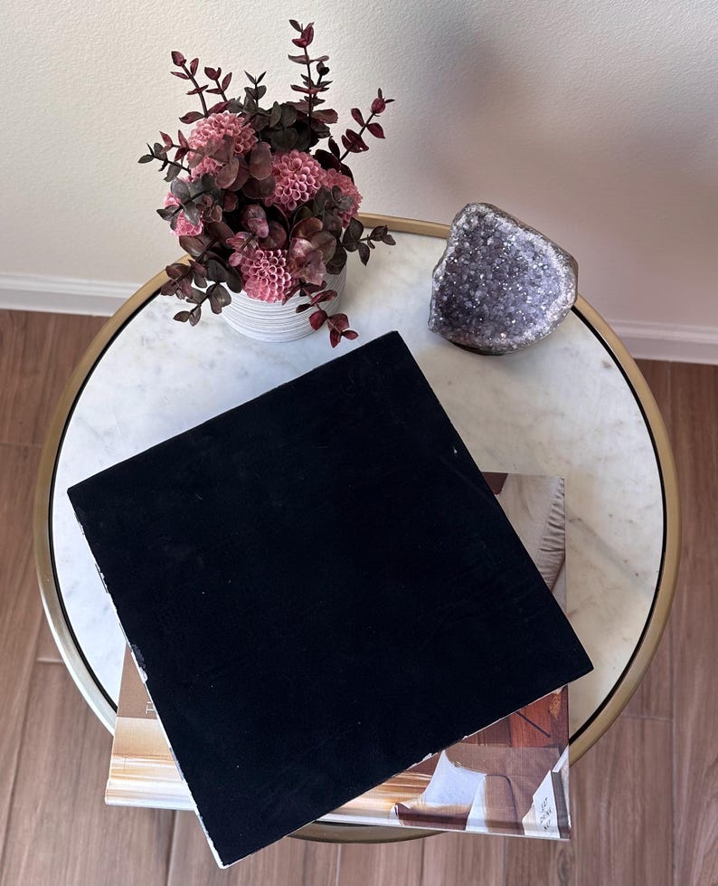 May include: A square black velvet display board resting on a book, accompanied by a white ribbed vase containing pink flowers with dark foliage and a jagged purple amethyst crystal geode, all arranged on a round white marble table with a gold rim.