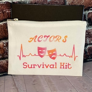 May include: A white canvas zipper pouch with the words "ACTORS Survival Kit" in orange and red lettering. The design includes two theater masks and a heartbeat line. A black box is visible behind the pouch.