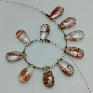 Puede incluir: Un hilo de piedras preciosas transparentes en forma de lágrima con inclusiones de color marrón rojizo. Las cuentas están ensartadas con alambre plateado y pequeñas cuentas doradas. Las piedras preciosas facetadas capturan la luz, mostrando sus patrones internos.