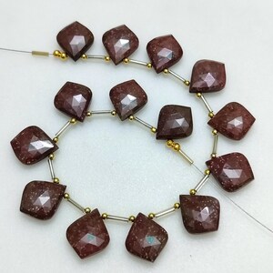 Puede incluir: Hebras de cuentas de piedras preciosas facetadas, de color marrón rojizo oscuro. Cada cuenta tiene una forma irregular única con múltiples facetas, ensartadas con alambre plateado y pequeñas cuentas doradas. Las cuentas se muestran sobre un fondo blanco.