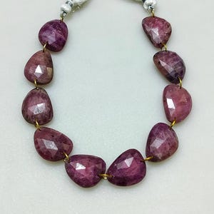 May include: A strand of faceted, oval-shaped, deep magenta gemstones. Each stone is connected with gold-colored wire and strung on a white cord. The gemstones have a polished, reflective surface.