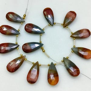 May include: Strand of faceted teardrop-shaped gemstones in shades of red, brown, and gray. The gemstones are strung together with gold-colored beads and silver-colored wire. The gemstones are approximately 1 inch in length.