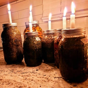 May include: A collection of glass mason jars filled with herbs and spices, each topped with a lit white candle. The jars are arranged on a countertop, with a warm, inviting glow from the flames.