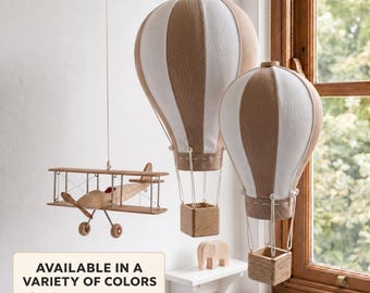 May include: Two hanging hot air balloon decorations and a biplane model, all featuring tan and white fabric panels. The balloons have small square wooden baskets suspended by white cords. A text box reads: AVAILABLE IN A VARIETY OF COLORS.