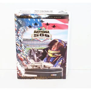 Puede incluir: Un programa de recuerdo para la carrera Daytona 500, con una foto de un piloto de carreras con casco y un modelo de coche de carreras dorado. El programa se titula "Daytona 500" con la fecha "15 de febrero de 2004".