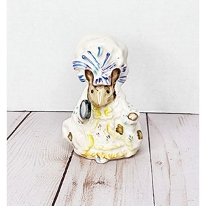 May include: A white ceramic figurine of a mouse wearing a blue and white hat and holding a magnifying glass. The mouse is wearing a white dress with floral details and is sitting on a wooden surface.