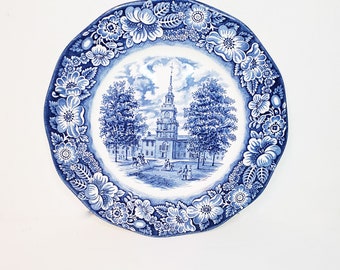 May include: Blue and white ceramic plate featuring a floral border and a central image of a building with a clock tower. The building is surrounded by trees and figures.