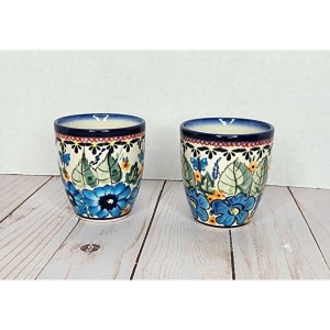 May include: Two ceramic mugs with a blue and white floral pattern. The mugs have a white background with blue flowers, green leaves, and yellow accents. The rim of the mugs is a dark blue color.