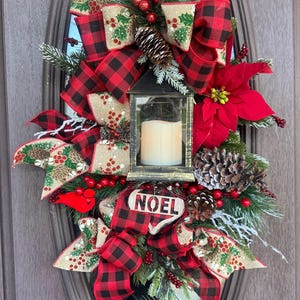 Pode incluir: Uma grinalda de Natal festiva com uma lanterna preta com uma vela. A grinalda apresenta laços xadrez vermelhos e pretos, pinhas, bagas vermelhas e um letreiro "NOEL". A grinalda é decorada com vegetação e uma flor de Natal vermelha.