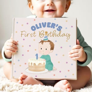 May include: A baby holds a book with the text "Oliver's First Birthday." The book cover features a cartoon illustration of a baby with a party hat next to a cake with a number one candle. The background is light pink with colourful polka dots.