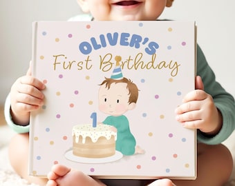 May include: A baby holds a book with the text "Oliver's First Birthday." The book cover features a cartoon illustration of a baby with a party hat next to a cake with a number one candle. The background is light pink with colorful polka dots.