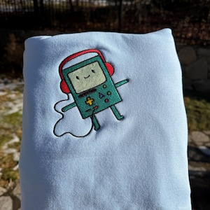 Puede incluir: Sudadera azul claro con un diseño bordado de un personaje verde y sonriente de consola de videojuegos retro con auriculares rojos. El personaje tiene brazos y piernas y está conectado a un cable blanco.