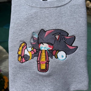 Könnte beinhalten: Ein graumeliertes Sweatshirt mit Rundhalsausschnitt und einer detaillierten Stickerei des Charakters Shadow the Hedgehog. Die Figur ist in einer liegenden Pose mit schwarzem Fell, roten Akzenten, gelben Schuhen und blauen Energieeffekten dargestellt.