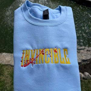 May include: A light blue crewneck sweatshirt featuring the word INVINCIBLE embroidered in yellow thread across the chest. The lettering is accented with a red embroidered splatter pattern that appears to cut diagonally across the text.