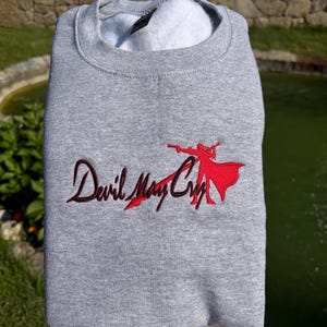 May include: A heather gray crewneck sweatshirt featuring an embroidered design on the chest. The embroidery includes the text Devil May Cry in black script lettering alongside a red silhouette of a character holding a gun and wearing a long coat.