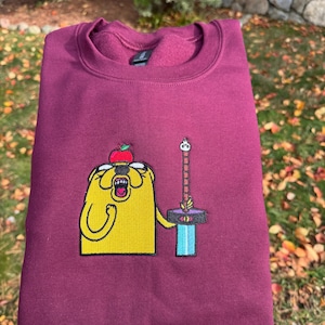 Op de afbeelding: Bordeauxkleurige crewneck sweatshirt met een geborduurd stripfiguur met een appel op zijn hoofd en een zwaard. Het personage is geel met zwarte contouren en houdt een zwaard vast met een blauwe en paarse pareerstang.