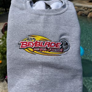 Puede incluir: Una sudadera gris jaspeado de cuello redondo con el logotipo bordado de Beyblade Metal Fusion en el pecho. El logo incluye letras rojas y blancas con un gráfico de llama amarilla y un emblema circular plateado que muestra una cabeza de lobo.