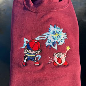 Peut inclure: Un sweat-shirt bordeaux avec des personnages de dessins animés brodés. Un personnage porte un chapeau rouge et bleu et un short bleu, avec une explosion d'énergie bleue et blanche. Un autre est une créature rose avec une baguette étoile.