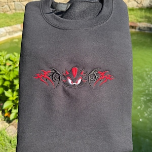 May include: A black crewneck sweatshirt featuring a central embroidered design of a stylised character head with red and black details, flanked by symmetrical red and black tribal-inspired patterns across the chest area of the garment.