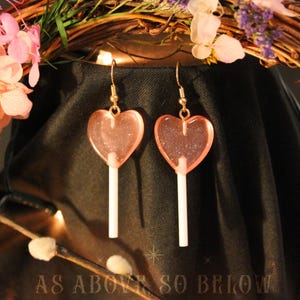 May include: Two heart-shaped earrings, designed to look like pink lollipops, with a glittery effect. They hang from gold-coloured hooks. The background is a dark fabric, with floral decorations and the text "AS ABOVE SO BELOW".