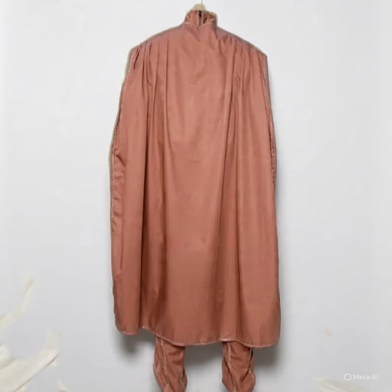 May include: A dusty rose-colored outfit, including a long cape and matching pants, hangs on a wooden hanger against a white wall. The cape has a high collar and a flowing design. The pants are visible at the bottom of the image.