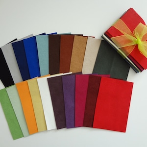 May include: A collection of colorful felt sheets in various shades, including black, blue, green, orange, and red. A stack of red felt sheets is tied with a yellow ribbon. The felt sheets are arranged on a white surface.