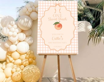 May include: A rectangular sign on a wooden easel featuring a light peach gingham pattern, a central scalloped frame, a peach illustration, and text reading ONE Sweet Peach, WELCOME TO Callie's FIRST BIRTHDAY PARTY, and 17.10.202X.