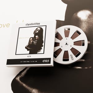 Könnte beinhalten: Eine weiße Box mit dem Text "OpenReelShop" und einem Schwarz-Weiß-Bild einer Frau. Eine Rolle braunes Band liegt neben der Box. Der Text "sade love deluxe" und "01/01" sind ebenfalls sichtbar.