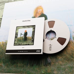 Könnte beinhalten: Ein weißes Box-Set mit dem Text "OpenReelShop" und "Loreena McKennitt" mit einem Foto einer Frau auf einem Feld. Neben dem Box-Set befindet sich ein Tonband. Der Hintergrund zeigt eine Person und den Text "PARALLEL DREAMS".