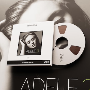 Puede incluir: Una caja blanca con el texto "OpenReelShop" y una imagen en blanco y negro de Adele. La caja está etiquetada como "ADELE 21" y está junto a una cinta de carrete. El carrete es blanco con cinta marrón.
