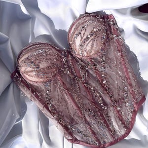 May include: A pink corset with intricate sequin and bead embellishments. The bustier features a sweetheart neckline and is made of sheer fabric with vertical lines of sequins. The edges are trimmed with a darker pink satin.