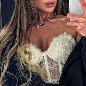 May include: Ivory lace corset with intricate floral patterns. The corset features a sweetheart neckline and a structured bodice. A silver chain necklace with a pendant is also visible. The image is a close-up shot.