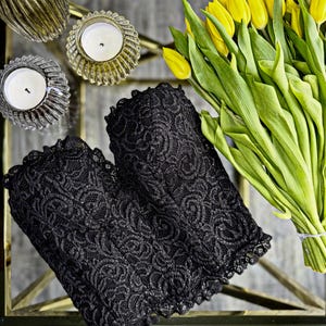 May include: Two black lace sleeves with a floral pattern are displayed on a glass surface. A bouquet of yellow tulips and two candle holders with lit candles are also visible. The lace has a scalloped edge.